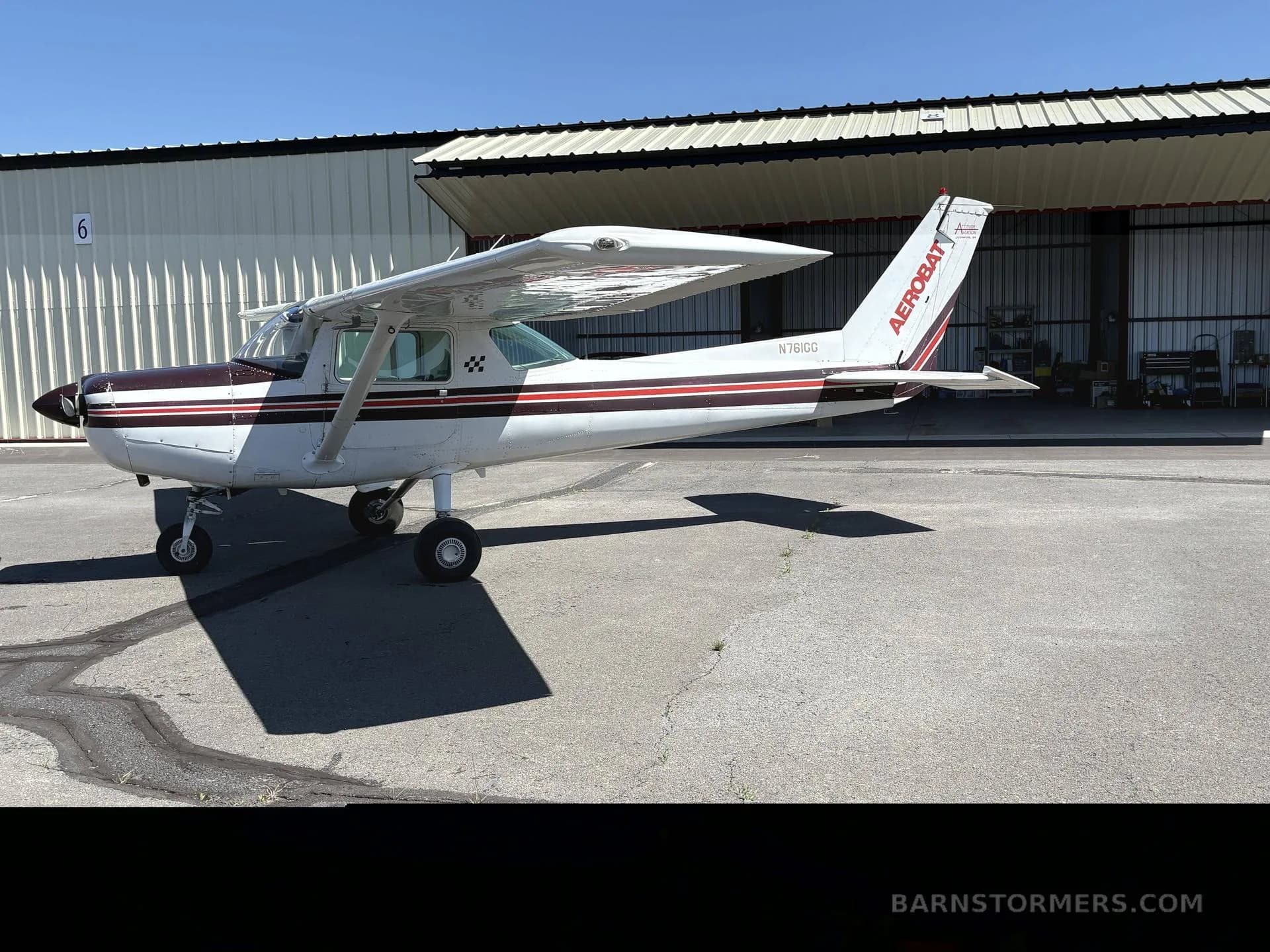 A white and maroon Cessna 150 Aerobat (N761GG) parked on an asphalt apron next to a hangar.