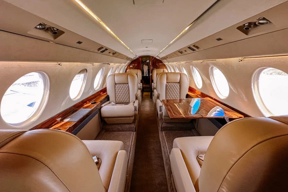 A view down the aisle of a luxurious aircraft cabin with tan leather seats and polished wood accents.