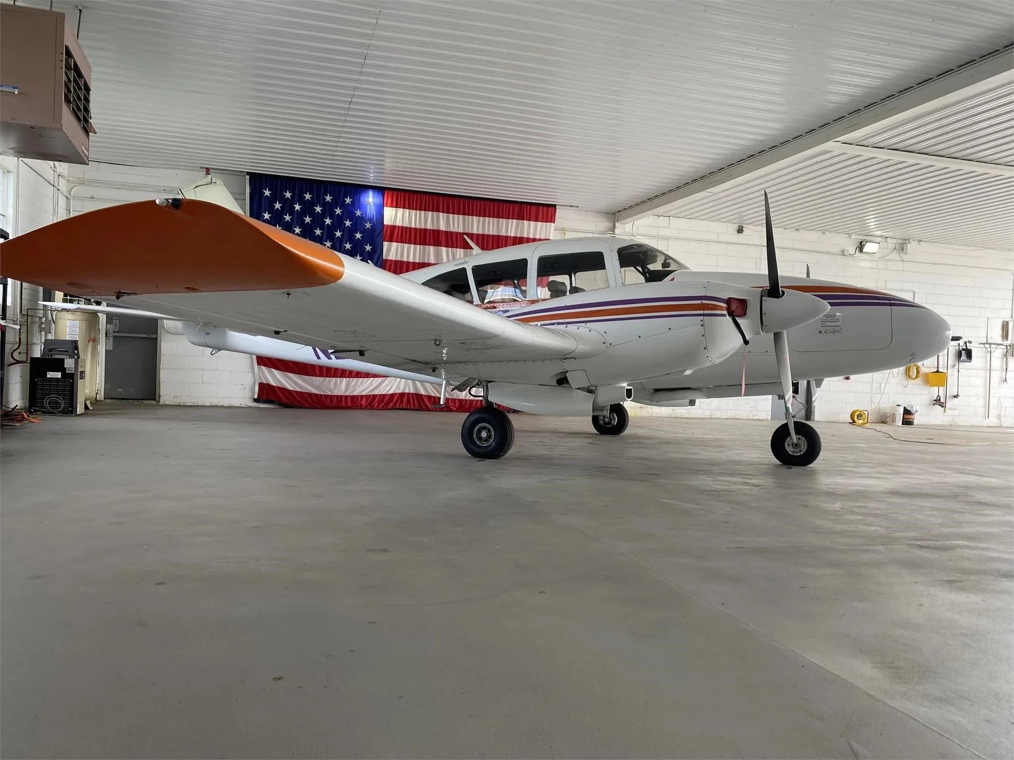 A white twin-engine Piper Aztec aircraft with orange and purple stripes parked in a hangar with an American flag in the background.
