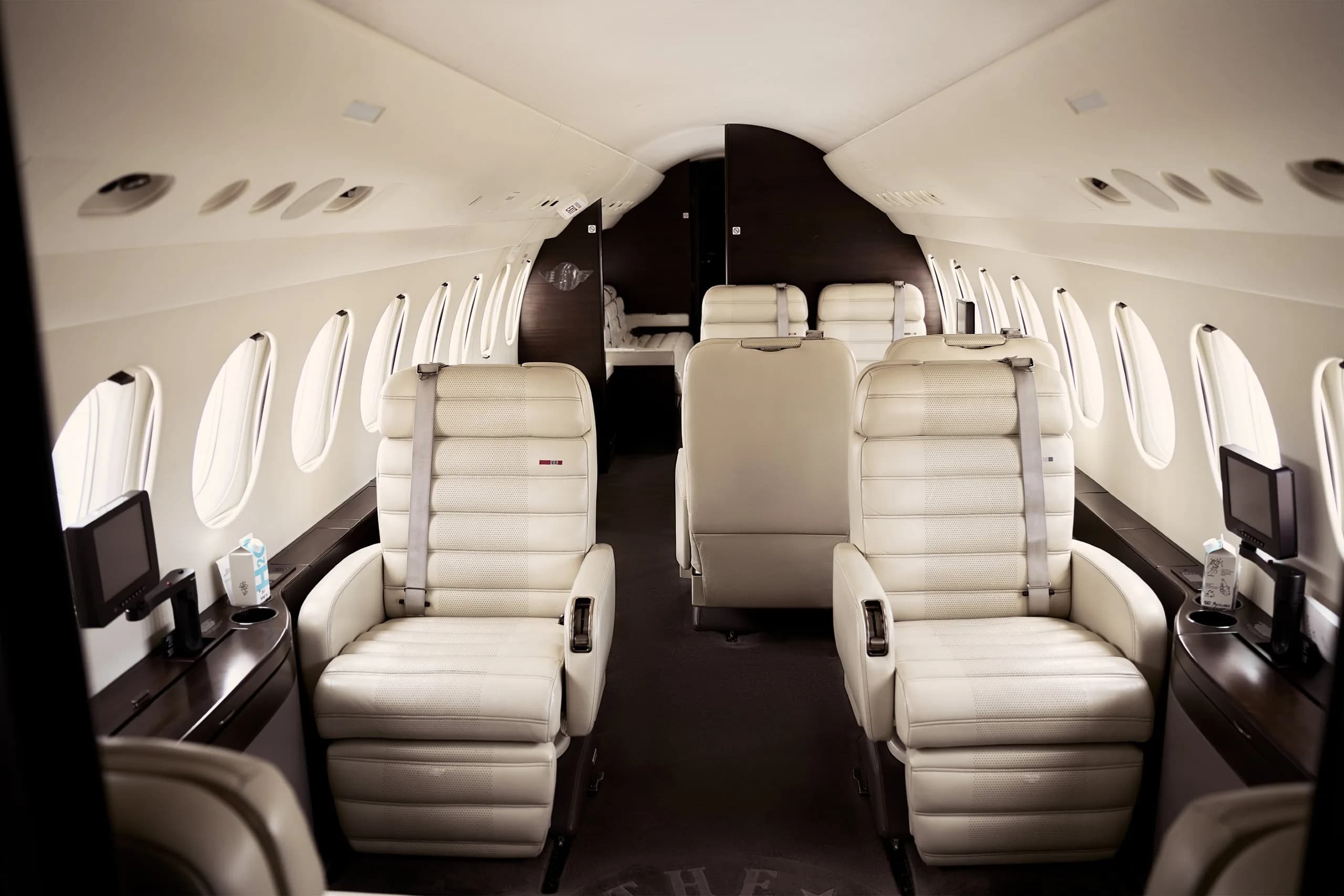 A wide view of a private jet cabin with multiple rows of cream leather seats and dark wood accents.
