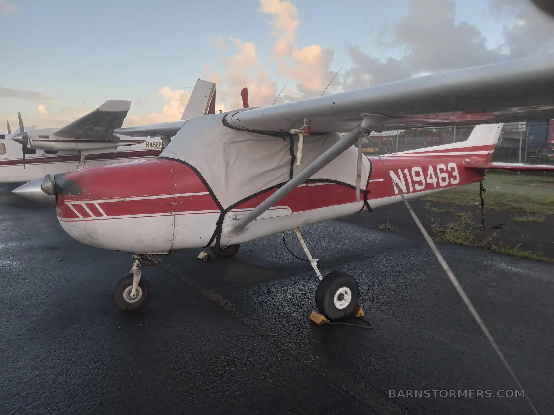A red and white Cessna 150 (N19463) parked on a wet tarmac with an engine cover.