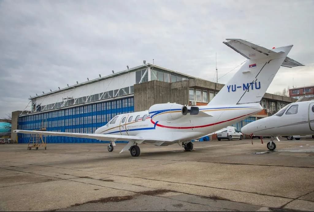 The tail and right side of a white business jet with "YU-MTU" registration, parked on an apron in front of a large building.