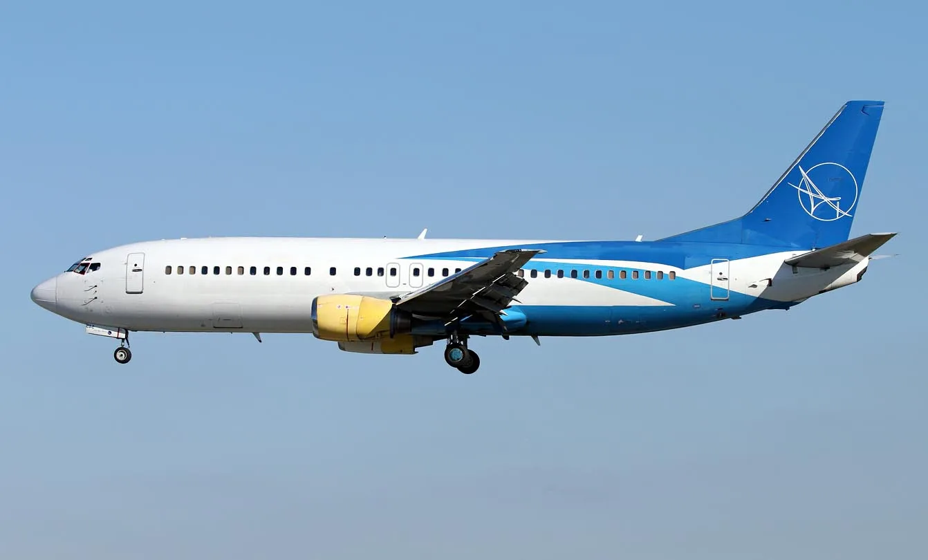 A white and blue passenger jet with a yellow engine nacelle is flying against a clear blue sky.