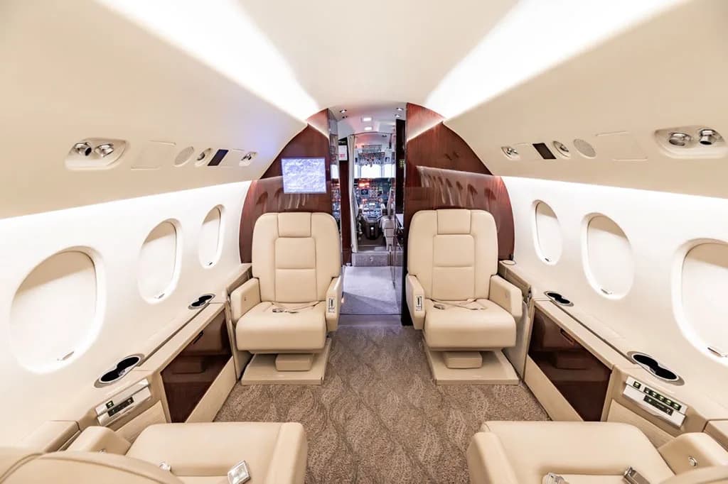The forward section of the business jet's luxurious interior cabin with beige seats and a screen visible.