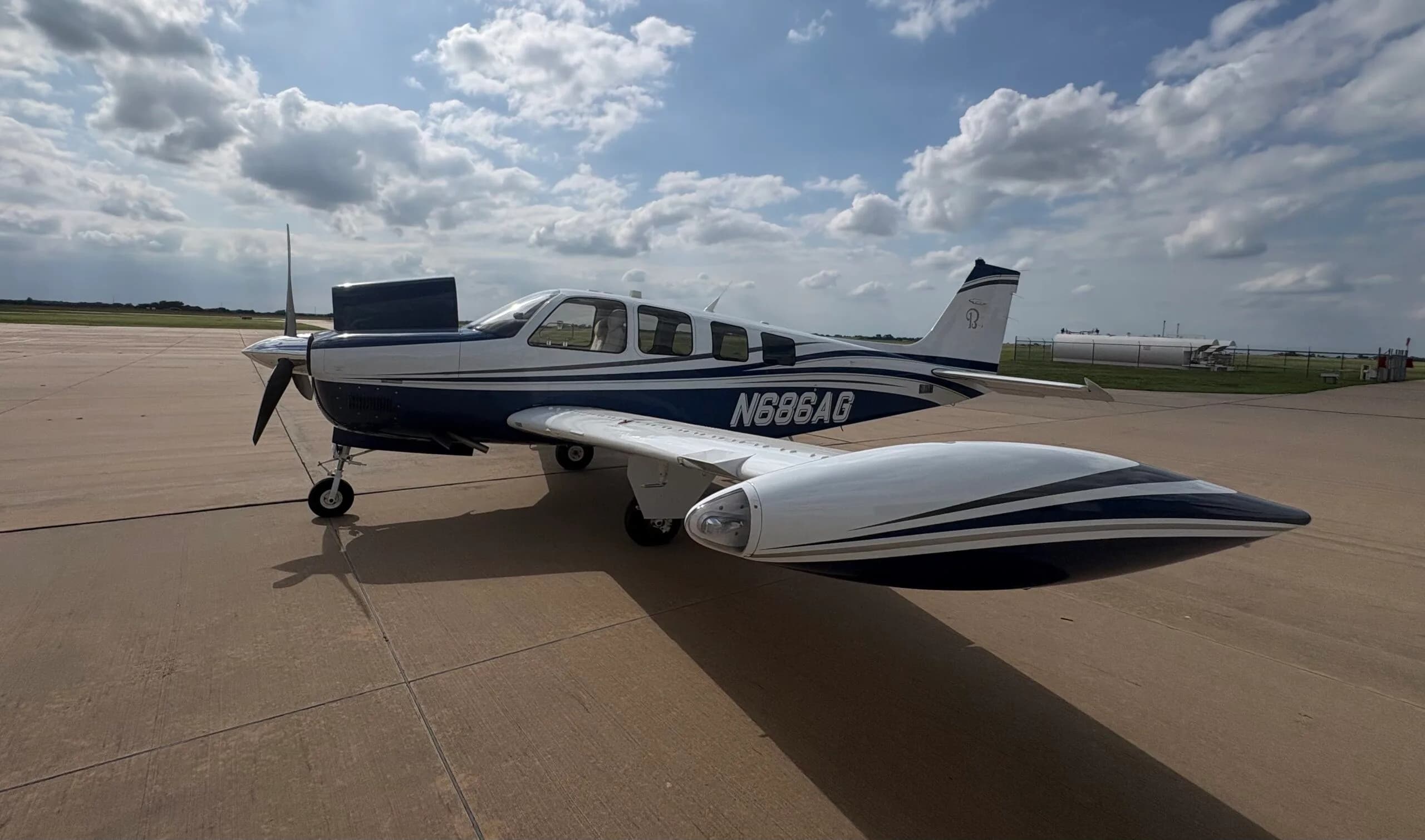 A three-quarter view of the white and blue Beechcraft Bonanza N686AG with its engine cowling open, parked on an airfield.