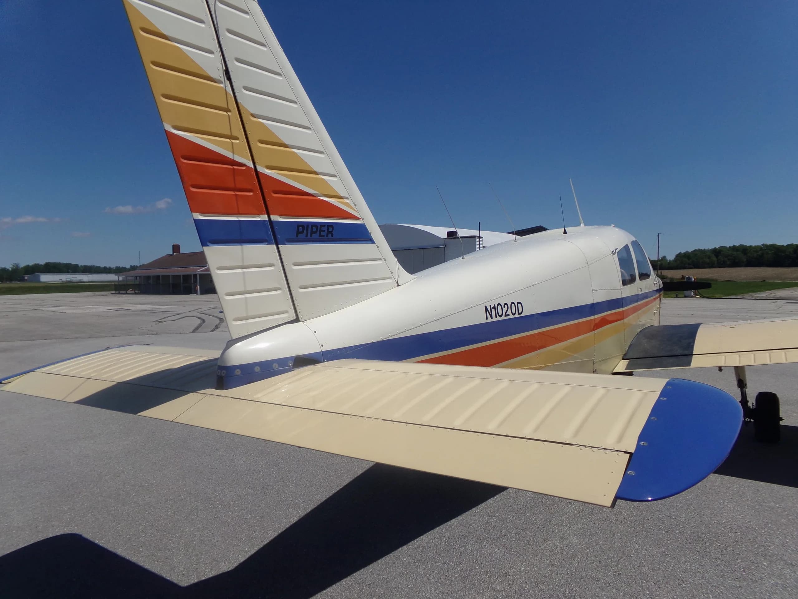 A rear-right view of a Piper aircraft, highlighting the tail with yellow, orange, and blue stripes and the "PIPER" logo.