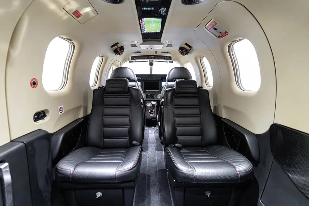 A view looking forward into a small aircraft cabin with four black leather seats and a screen in the ceiling.