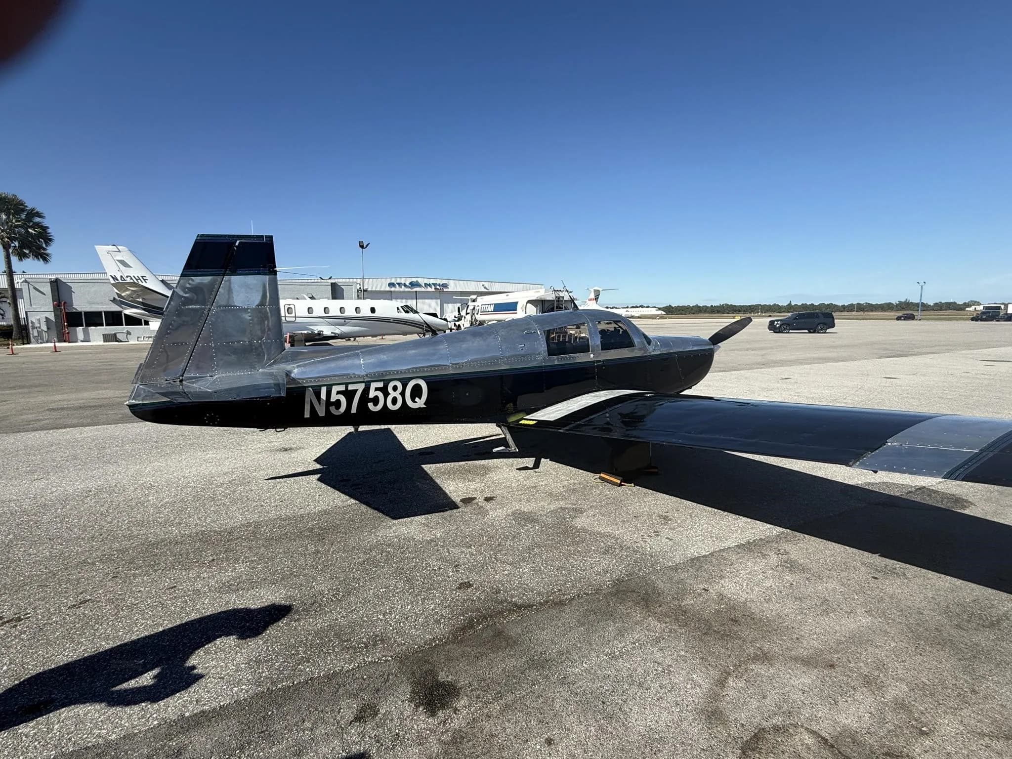 A black and silver Mooney M20 aircraft, N5758Q, parked on an asphalt tarmac with hangars and palm trees in the background.