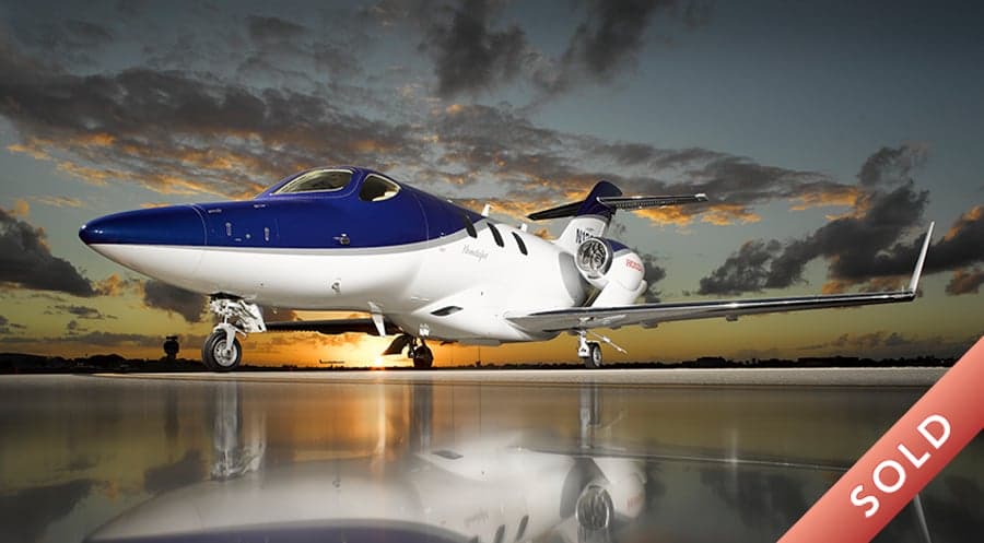 A HondaJet with a blue and white paint scheme is parked on the tarmac at sunset.