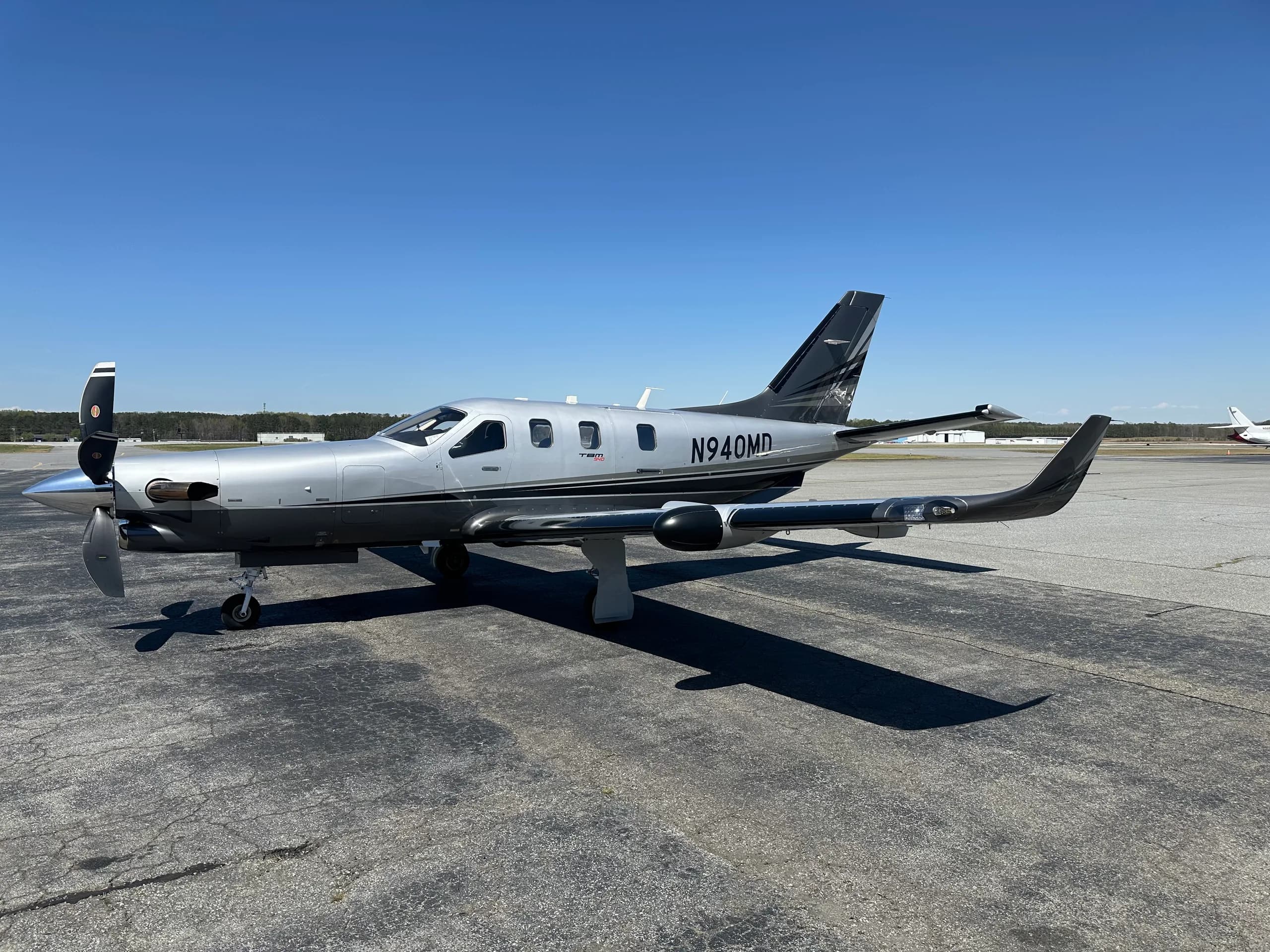 A full side view of a silver and black TBM 940 aircraft with registration N940MD, parked on an airport tarmac.