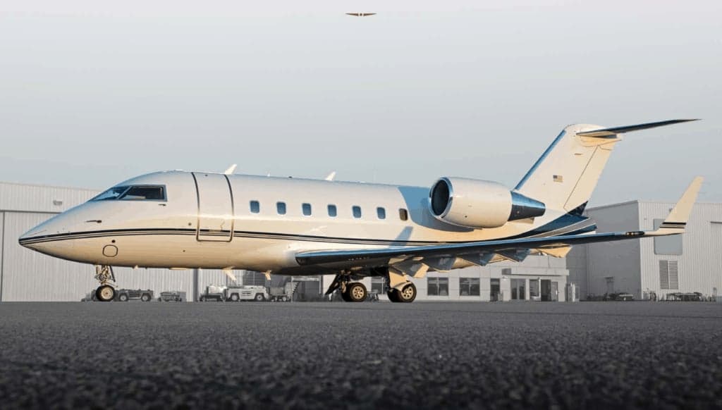 A white business jet with dark blue stripes is parked on an airport tarmac with hangars in the background.