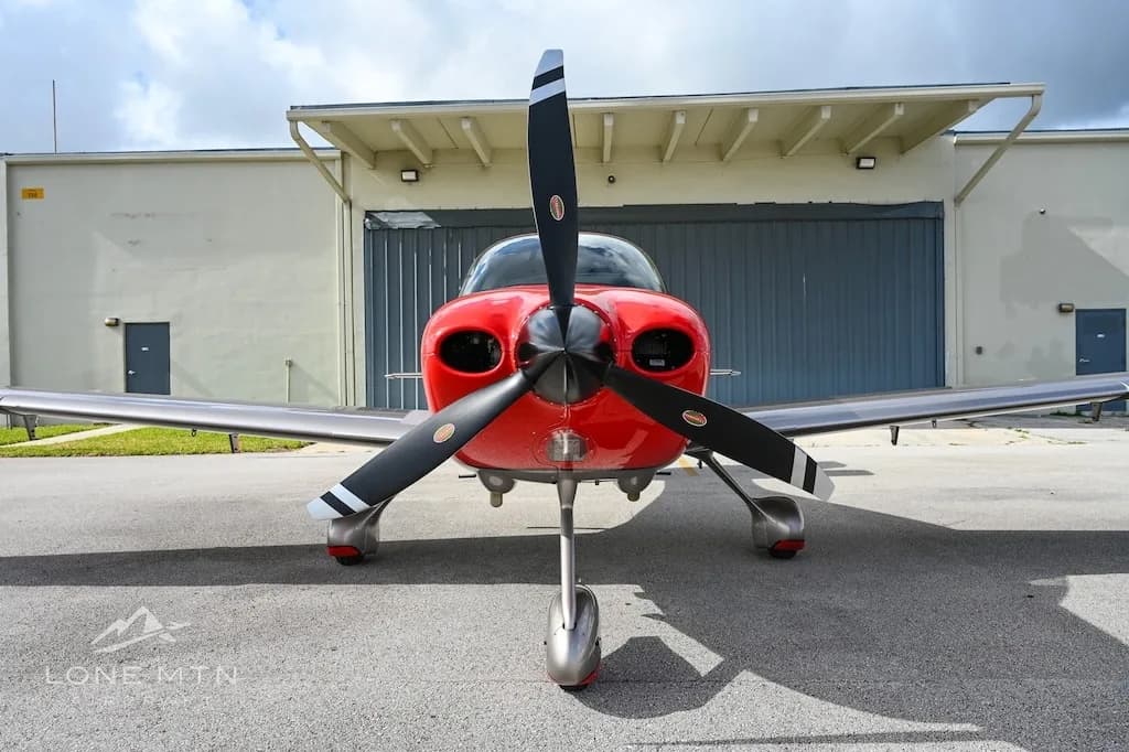 A head-on view of a red and silver Cirrus SR22 aircraft with a three-bladed propeller, parked in front of a hangar.