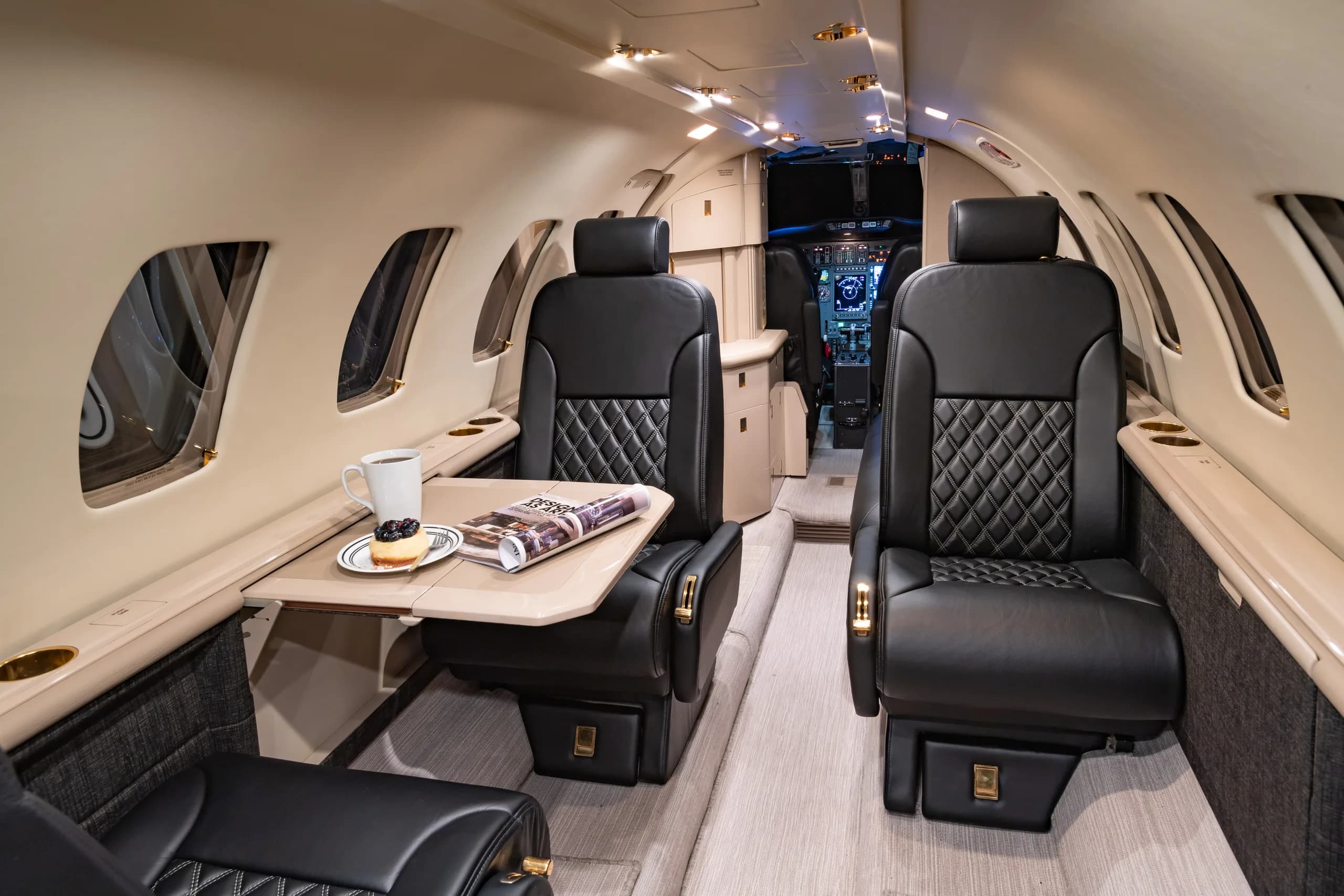 A close-up view of the business jet's cabin interior, featuring black quilted leather seats, a table with a magazine, coffee, and a pastry, and a partial view of the cockpit.