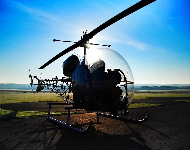 A vintage helicopter with a bubble canopy is silhouetted against a bright sunrise on an airfield.