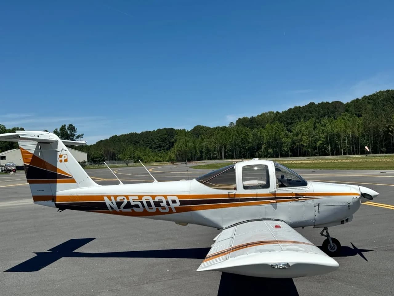 A white Piper PA-38 Tomahawk aircraft with orange and black stripes, seen from the right side on an airfield.