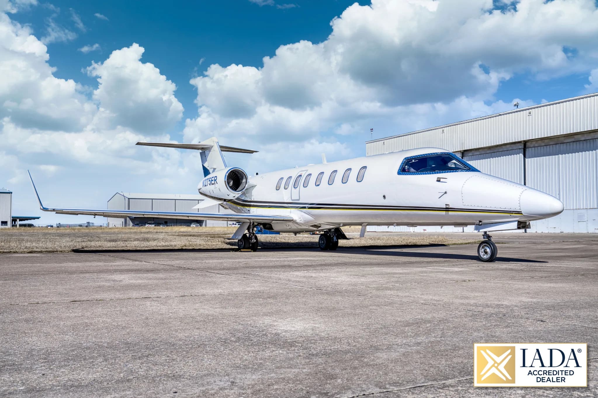 A three-quarter front view of a white Learjet 60 (N275ER) with a black and yellow stripe, parked on a tarmac in front of hangars.