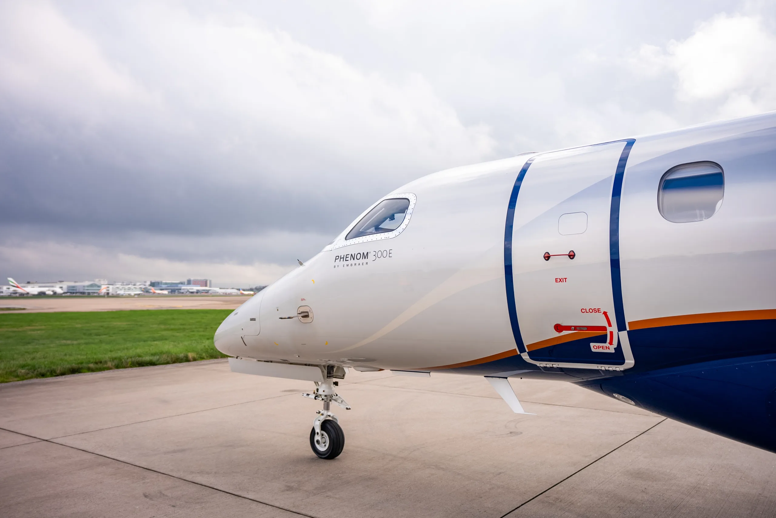 A close-up side view of the front fuselage and entry door of a white and blue Embraer Phenom 300E business jet on an airfield.