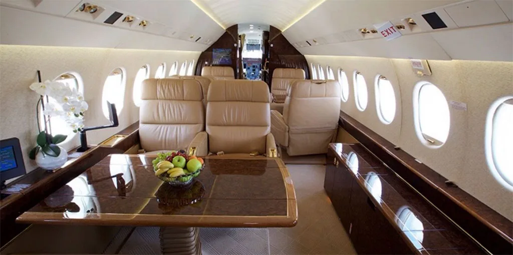 A wide view of the private jet cabin, featuring tan leather seats, a large polished wood table with fruit, and an orchid.