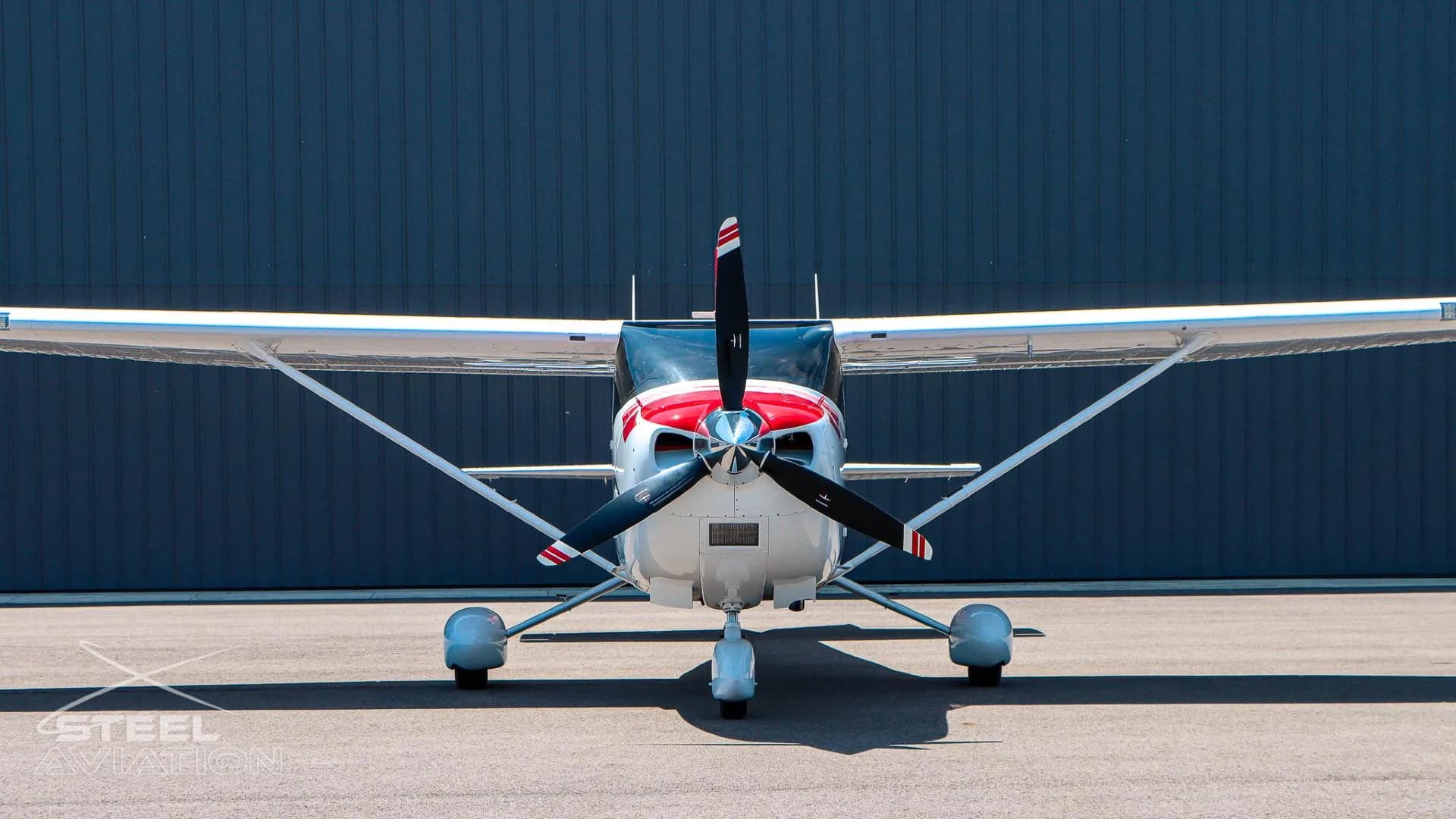 A white Cessna T182T Skylane TC with red and grey stripes is seen from the front, parked on an asphalt tarmac in front of a dark hangar.