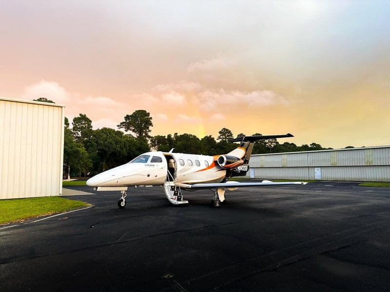 A Phenom 100 aircraft with registration N843SG is parked on the tarmac with its door open at sunset.