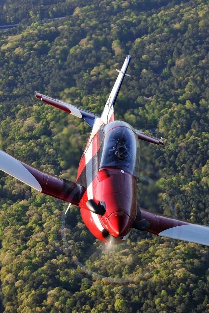 A red and white single-engine aircraft with a bubble canopy flies directly towards the viewer over a forest.