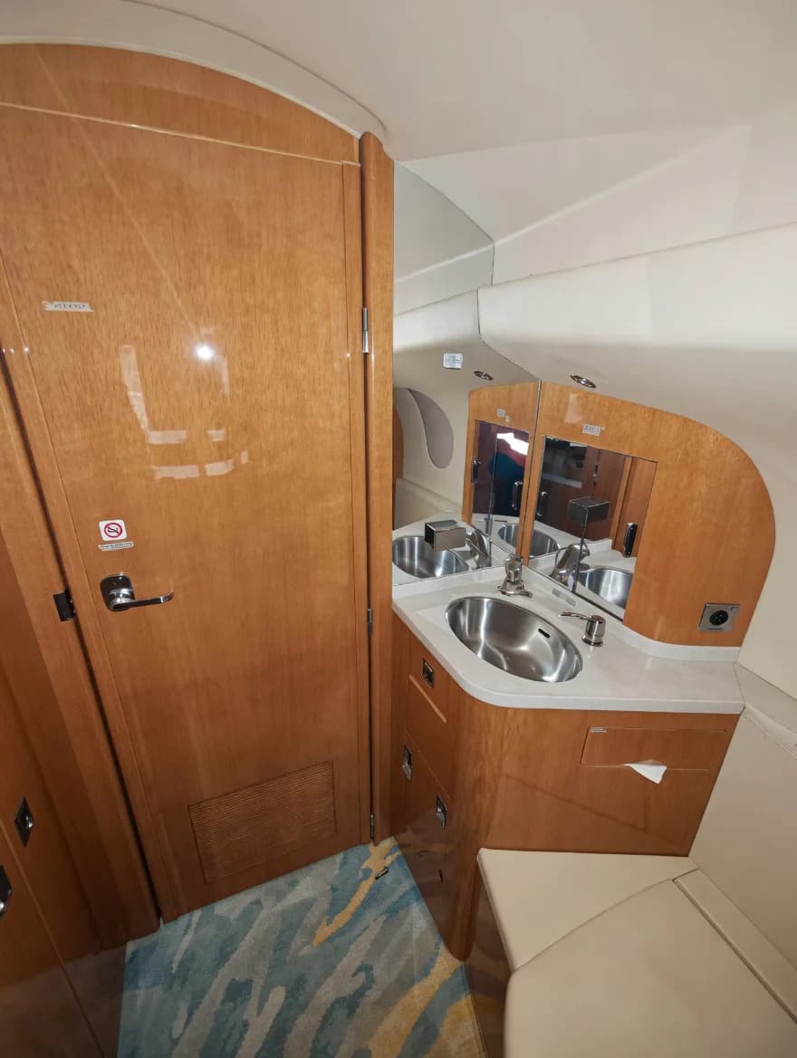 An aircraft lavatory with a wooden door, a vanity with a stainless steel sink, and a mirror.