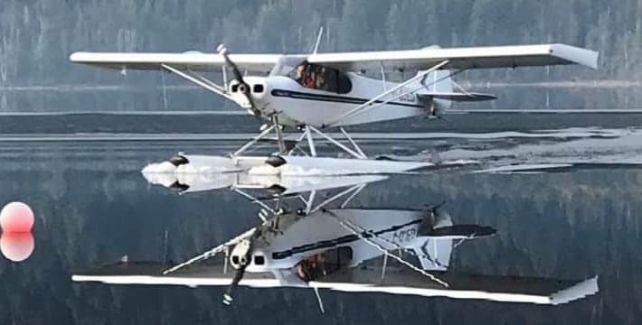 A white and blue Super Cub floatplane on the water with a reflection.