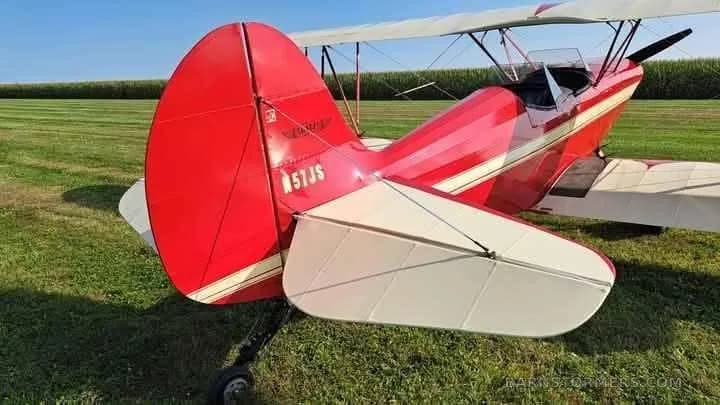 Rear view of a red biplane with white stripes and tail number N57JS, parked on a grassy field next to a cornfield.