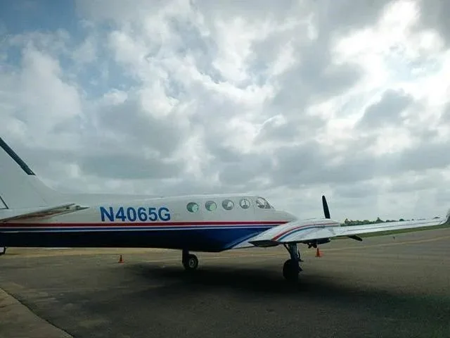 A side view of a white and blue Cessna 340 twin-engine aircraft with red stripes on an airfield under a cloudy sky.