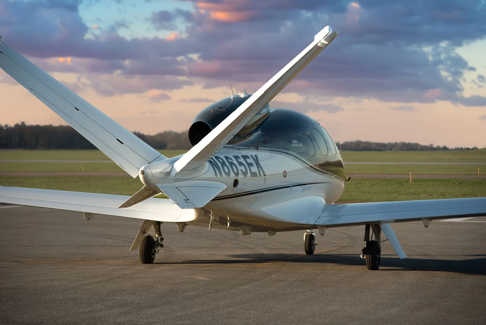 A rear three-quarter view of a Cirrus Vision Jet (N865EK) with a dark grey and white paint scheme parked on an airfield.