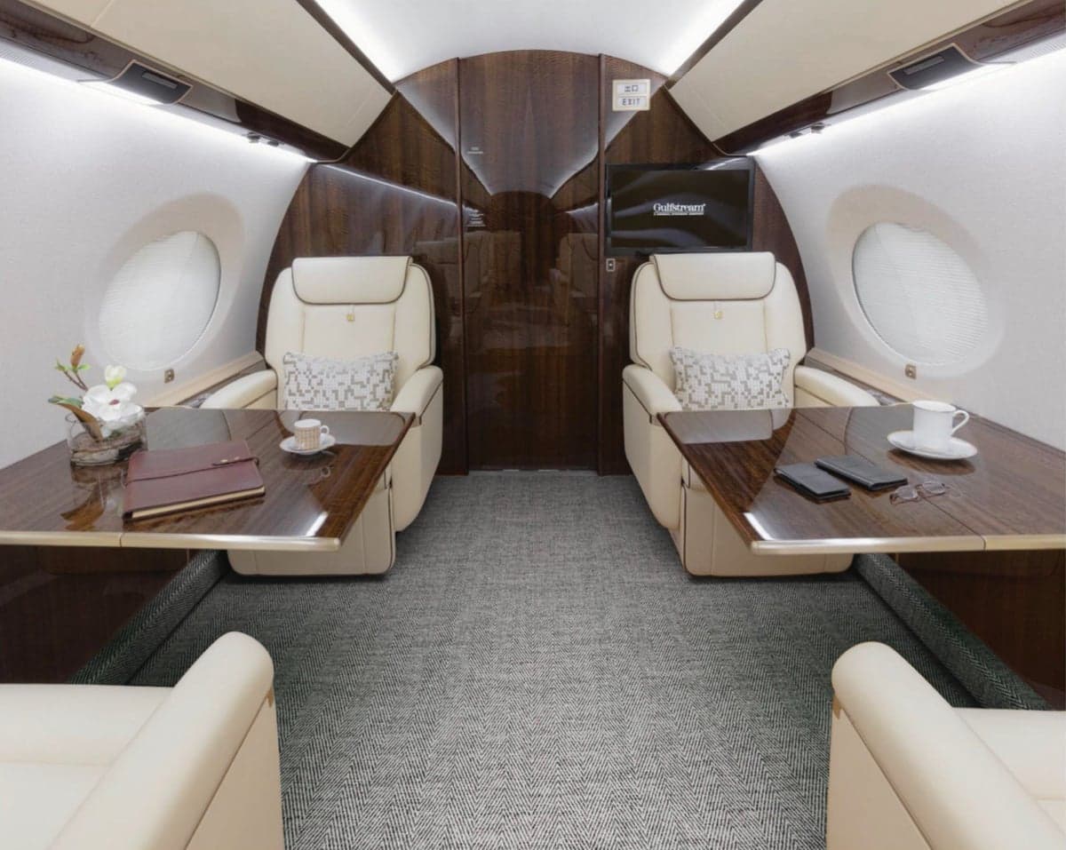 Aircraft cabin with cream-colored leather seats, tables, and a TV screen.