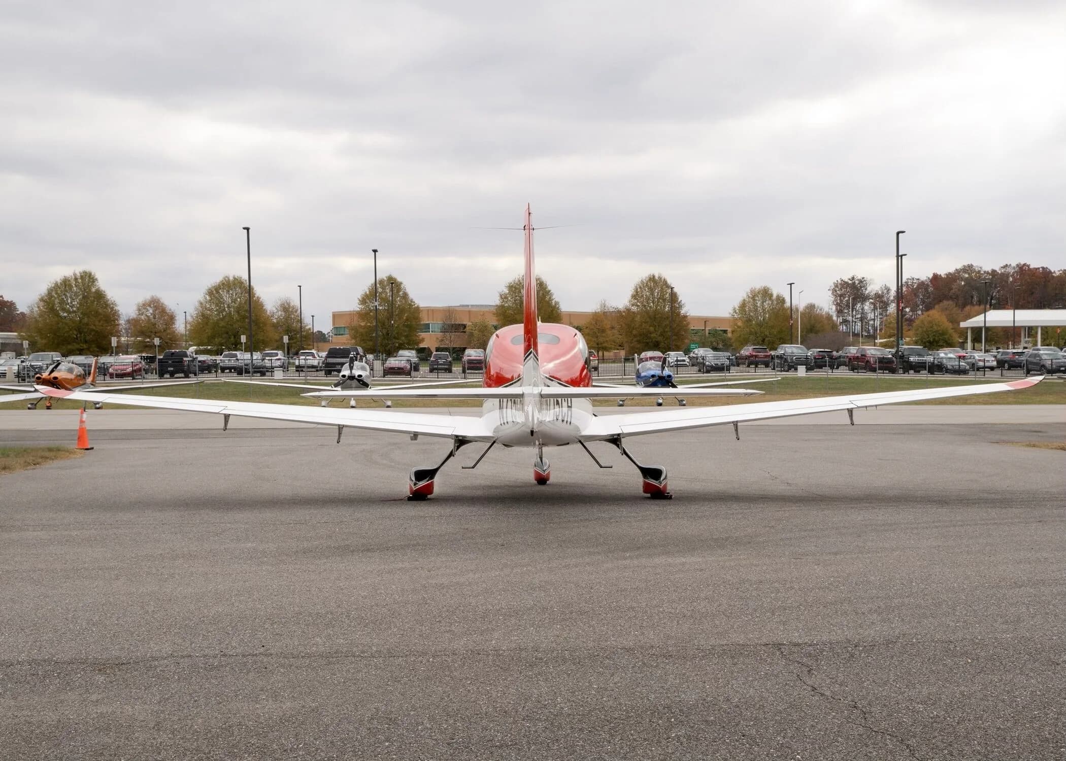 Rear view of a red and white Cirrus SR22 aircraft parked on an airfield with a parking lot in the background.