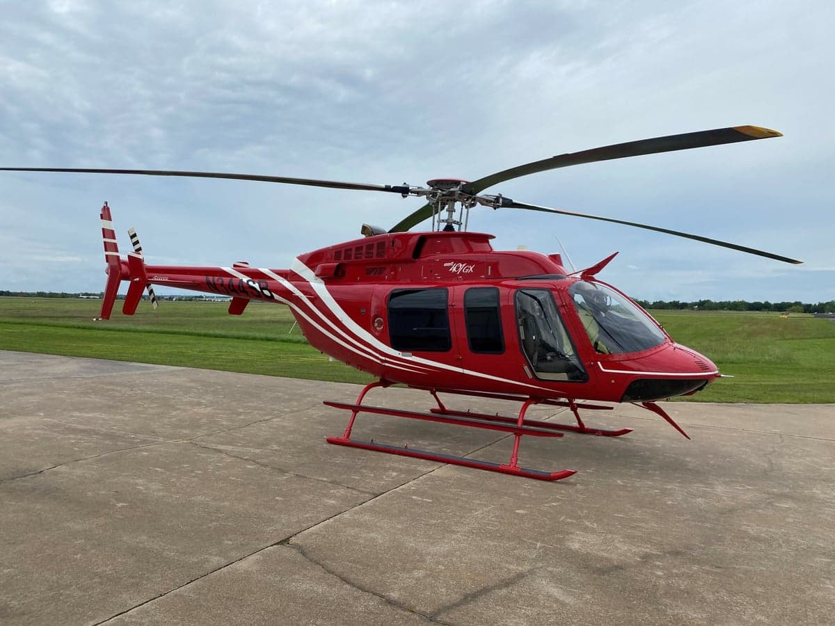 A red Bell 407GX helicopter with white stripes on its tail and fuselage, parked on a concrete pad at an airfield under a cloudy sky.