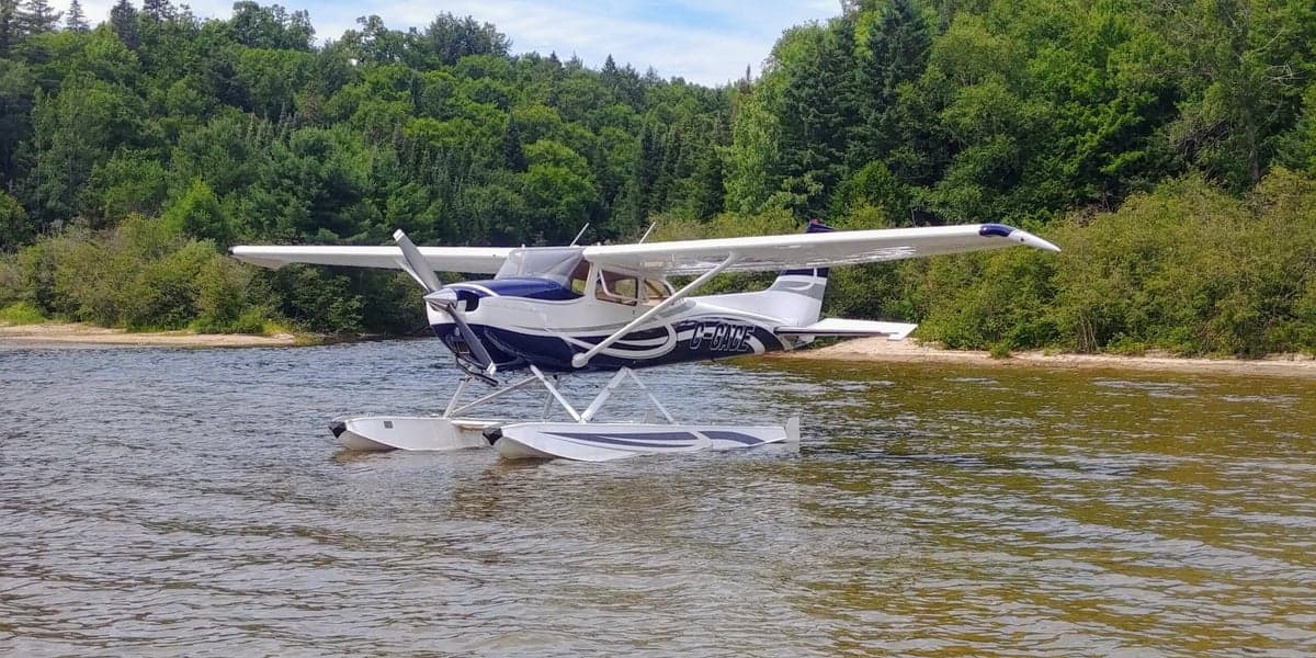 A Cessna floatplane sits in the water near a wooded shoreline.