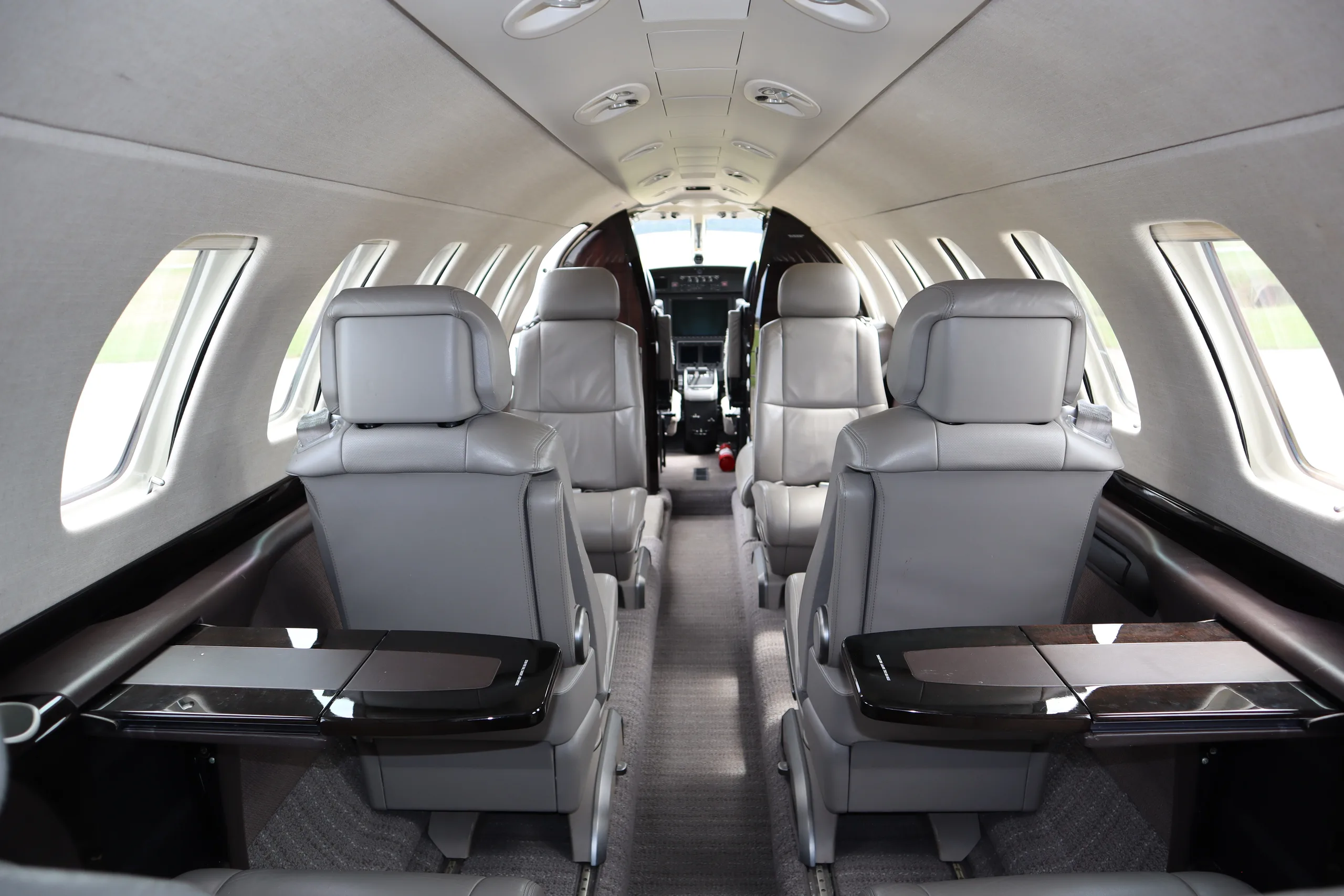 The interior cabin of a business jet, showing grey leather seats with fold-out tables, looking towards the cockpit.