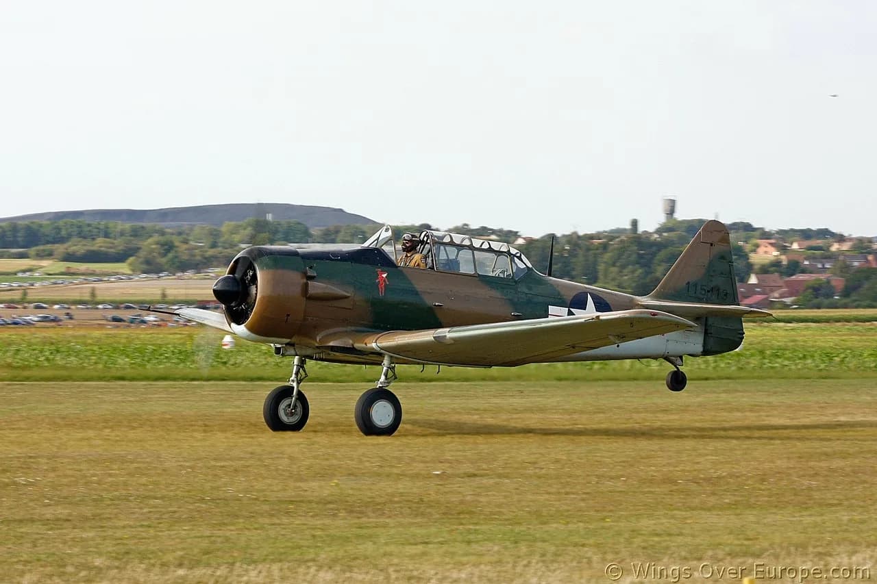 A North American T-6 Texan aircraft, F-AZAU, in camouflage livery, taxiing on a grassy airfield with a pilot visible in the cockpit.