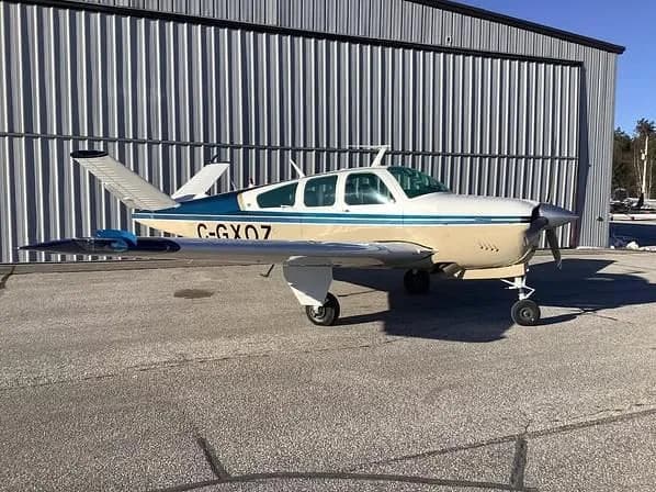 A beige and blue single-engine aircraft with registration C-GXOZ is parked on an asphalt apron next to a corrugated metal hangar.