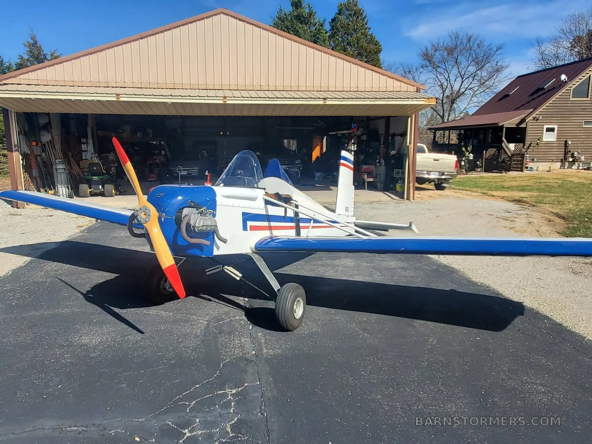 A white and blue single-engine aircraft with a wooden propeller is parked in front of an open hangar on a sunny day.