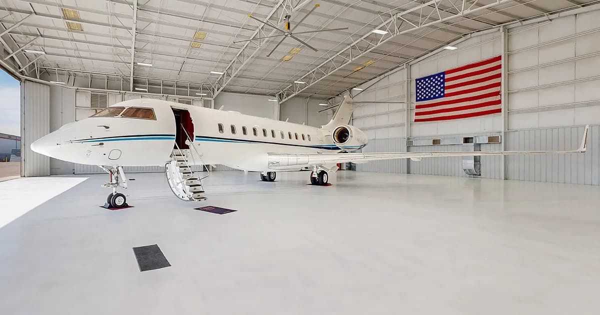 A white business jet with blue stripes parked in a brightly lit hangar with an American flag on the wall.