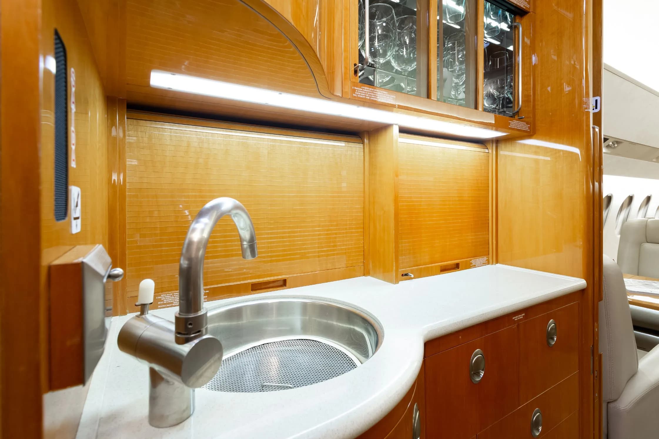 The galley area of a private jet, featuring a stainless steel sink, white countertop, and polished wooden cabinetry with glassware.