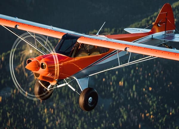 An orange and white Carbon Cub aircraft in flight over a forest.