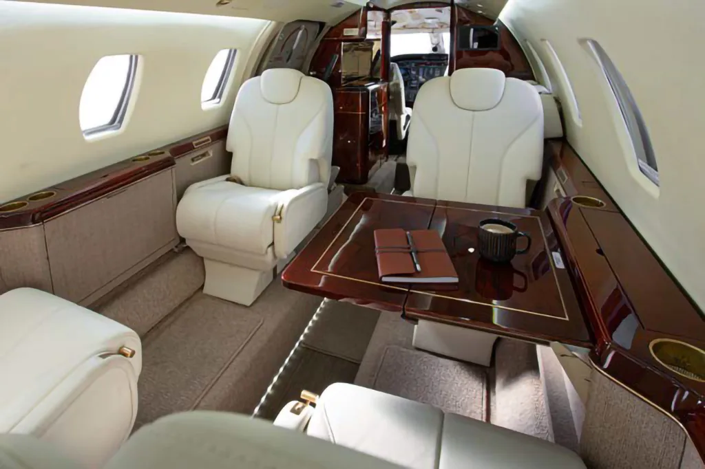 Wide view of the private jet cabin interior, featuring cream leather seating, a wooden table, and plush carpeting.