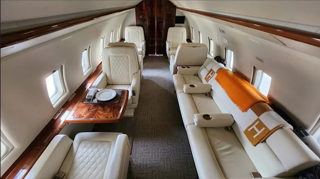 The luxurious aircraft cabin features cream leather seating, a polished wooden table set for dining, and an orange blanket on a sofa.