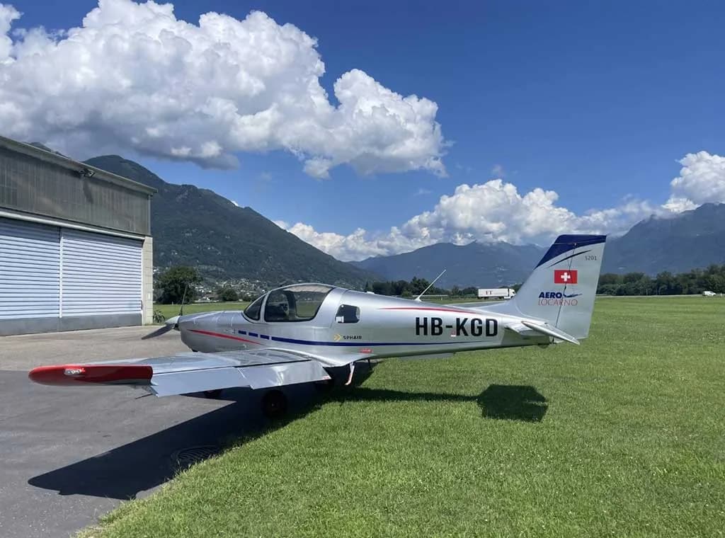 A silver Sonaca 200 aircraft (HB-KGD) is parked on a tarmac next to a hangar with mountains under a blue sky.