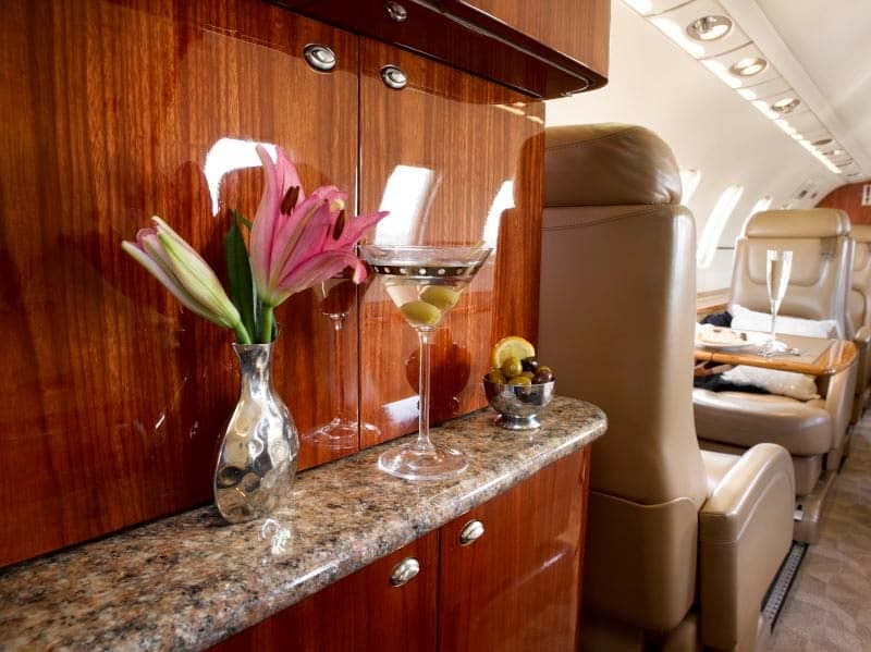 The aircraft cabin interior, showing a polished wood cabinet, granite countertop with flowers and drinks, and tan leather seats.
