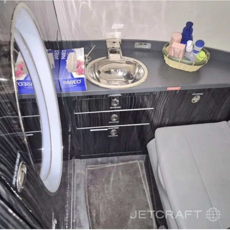 The lavatory area of a private jet, featuring a chrome sink, dark wood-grain cabinetry, and a lighted mirror.