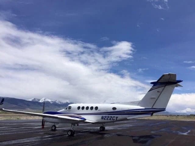A white and blue Beechcraft King Air with registration N222CY parked on a wet tarmac with mountains in the background.