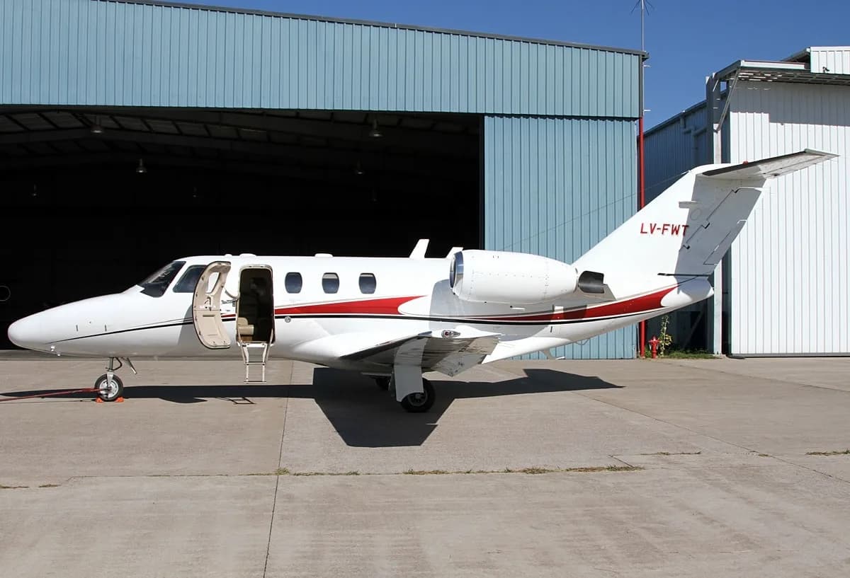 A white Cessna CitationJet with red and black stripes, registration LV-FWT, parked on an tarmac in front of a hangar with its main door open.