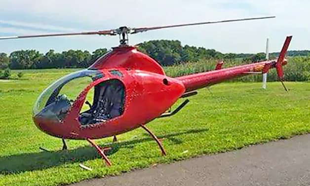 A red RotorWay Exec helicopter is parked on a grassy area next to a paved path.