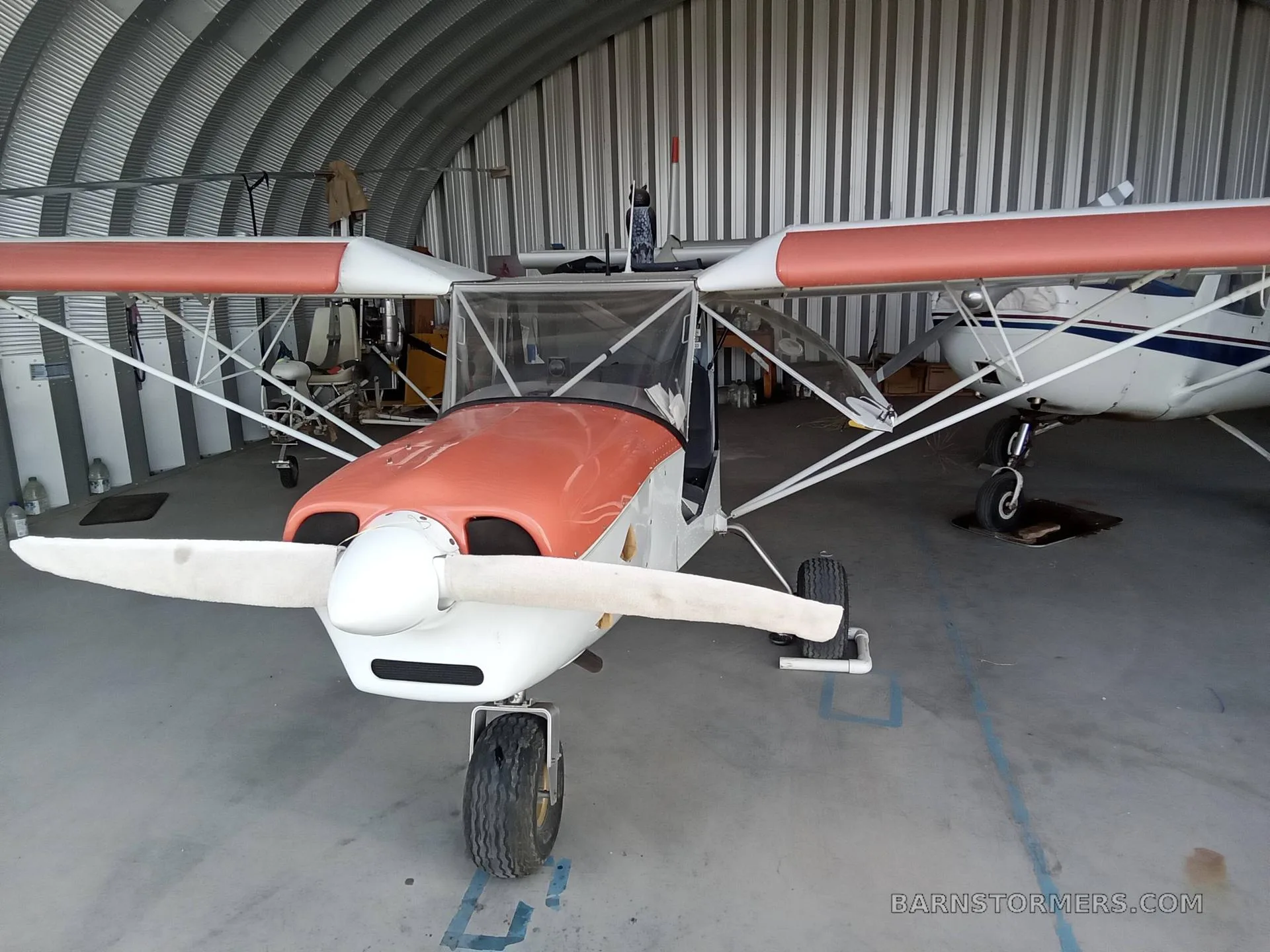 A light sport aircraft with an orange and white paint scheme is parked head-on inside a metal hangar, showing its propeller.